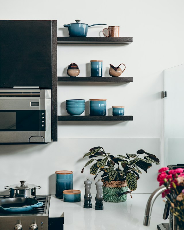 Teal ceramic pans, canisters and bowls on dark open shelving in a bright kitchen